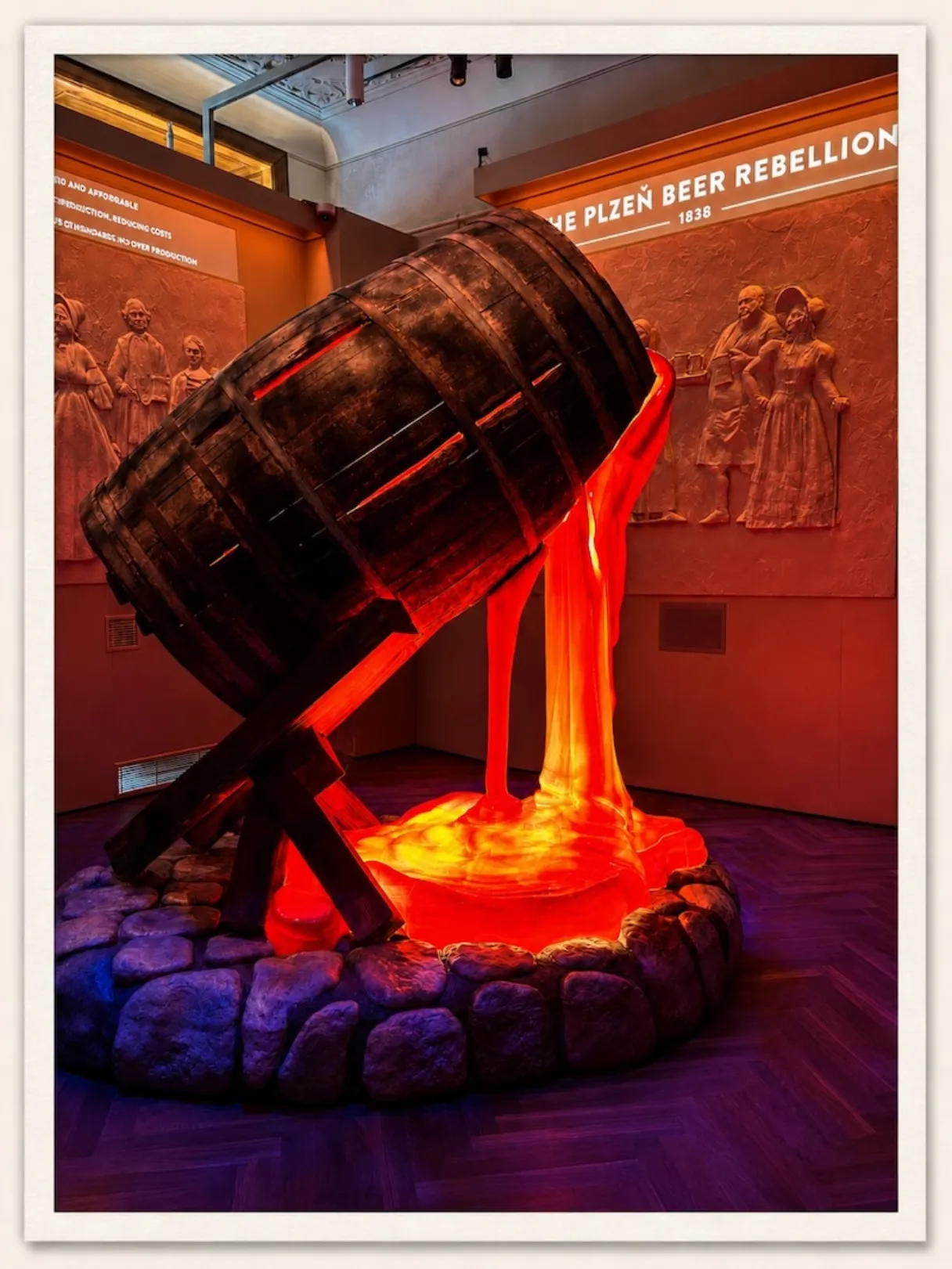 Photo from the Pilsner Urquell Experience in Prague, showing a large wooden beer barrel installation with glowing beer pouring into a stone base.